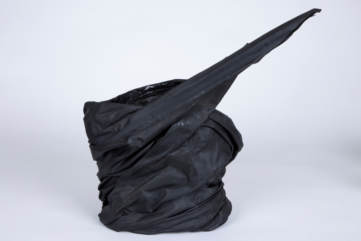 Kroppssmykke av svart silke som er drapert i en korsettlignende form og avstivet. Smykket er åpen foran, men drapert litt over hverandre. Endene er avsluttet i en spiss. Skumgummibånd i midjen på innsiden.