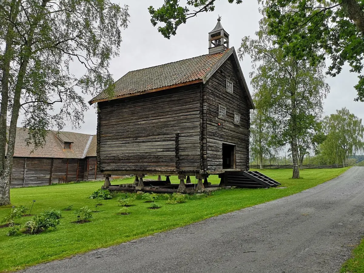 Bildet viser et stort stabbur. Det er sommer og gresset er grønt. Bak stabburet er det en stor låve.