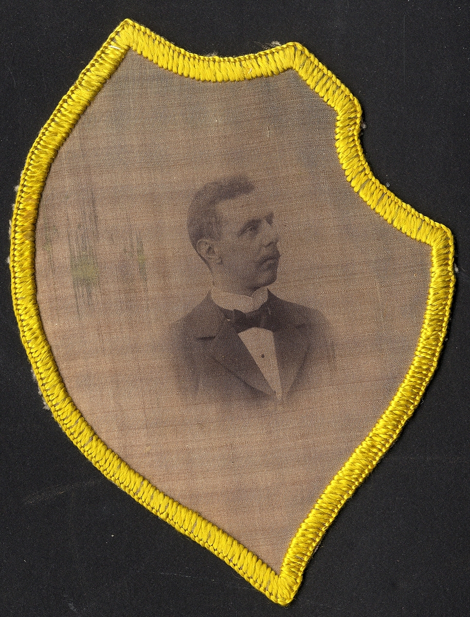 "Vapensköld" av sidenliknande tyg i grått med tryckt foto av en man i profil.
Bröstbild. 
Kan ev. vara: Ernst Ossian Ljunggren (1875-1942).