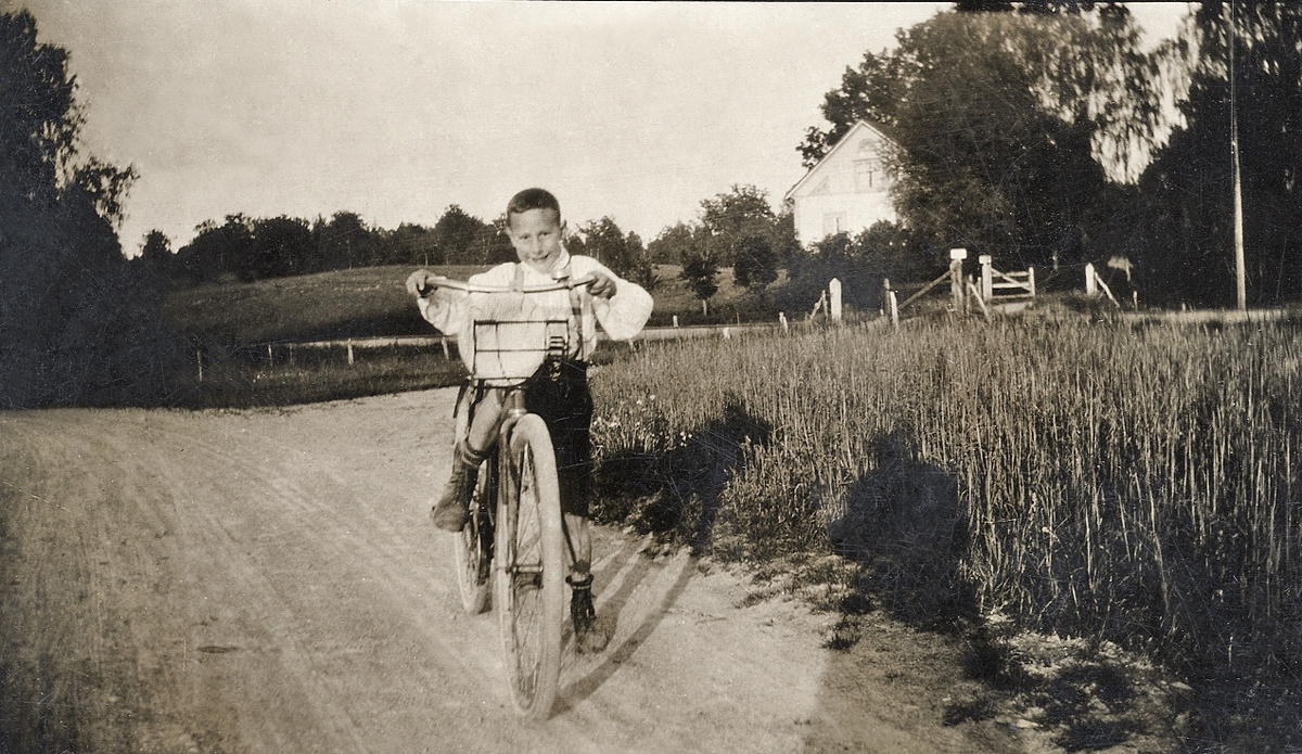 En glad pojke med (lite för stor) cykel försöker cykla på en grusväg. Till höger skymtar ett bostadshus i en trädgård.
