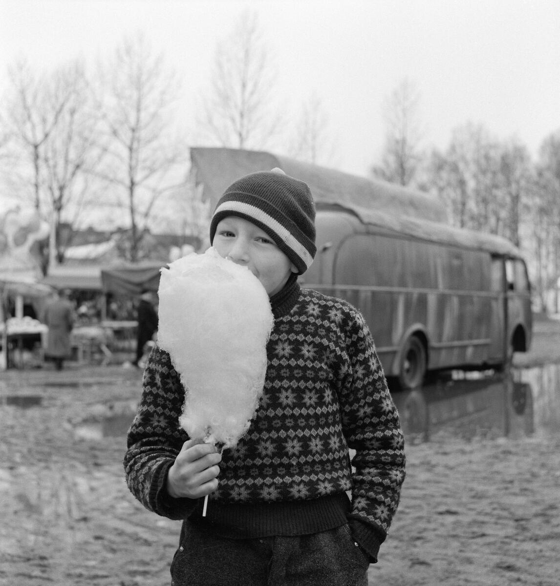 Svarthvitbilde av Gutt som spiser sukkerspinn. Har har på seg strikkeklær og lue med hvit bord. I bakgrunnen står en gammel buss