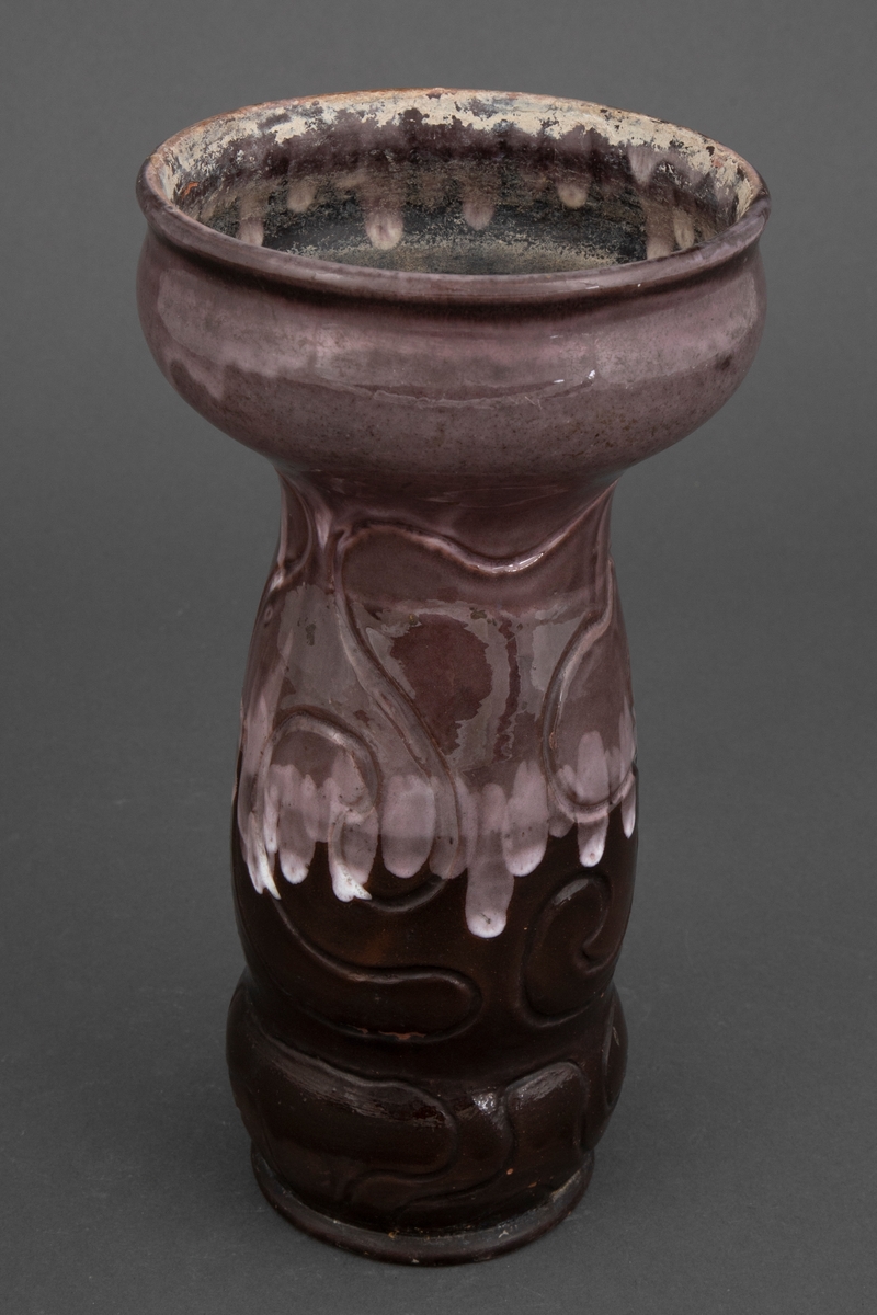 Vase i rødt gods med innrissede linjer som slynger seg rundt korpus. Nederste del av er glasert med blank glasur i brun, mens den øverste delen er dekket av brunfiolett rennende glasur med hvite partier.