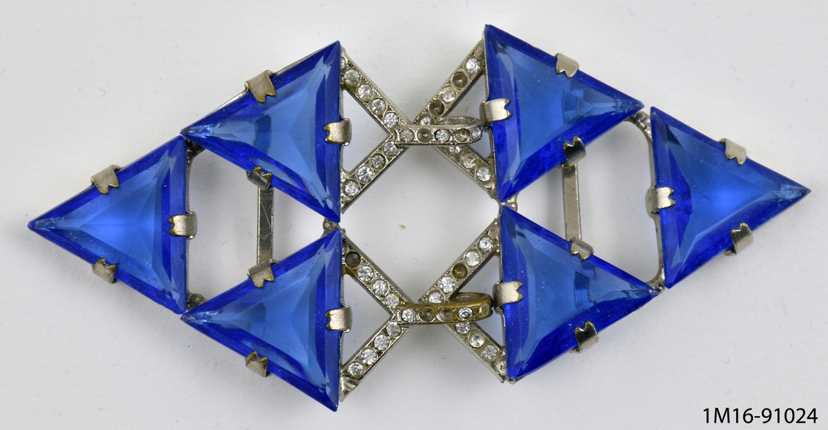 Skärpspänne av metall med glaspärlor, två delar. Sex stycken triangelformade blå glaspärlor fästade mot metall. Prismärkning: N.a.g. kr. 12:50.
