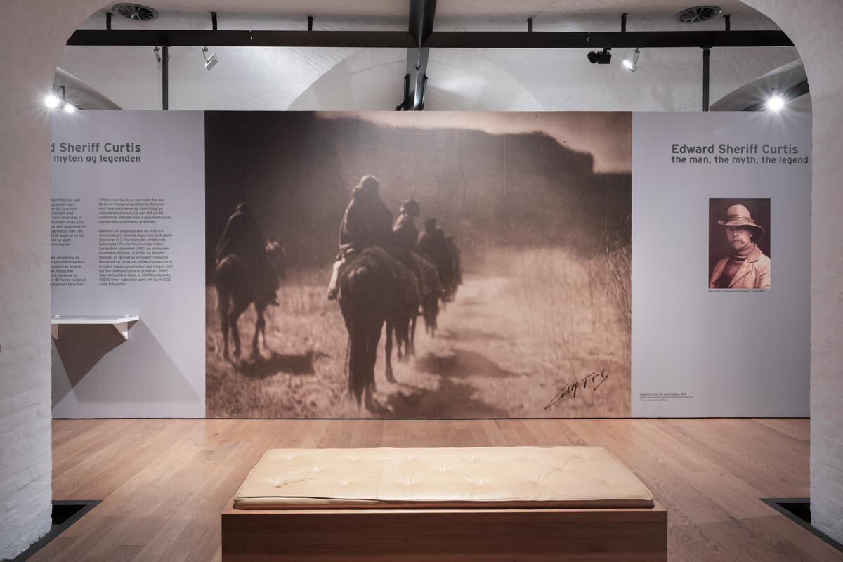 Dokumentasjonsfoto fra utstillingen Edward S. Curtis, mannen, myten og legenden. Bildet viser et av utstillingsrommene, på veggen er det et stort bilde av noen indianere som rir over et landskap med fjell rundt.  (foto: Ana Gonçalves/Preus museum)