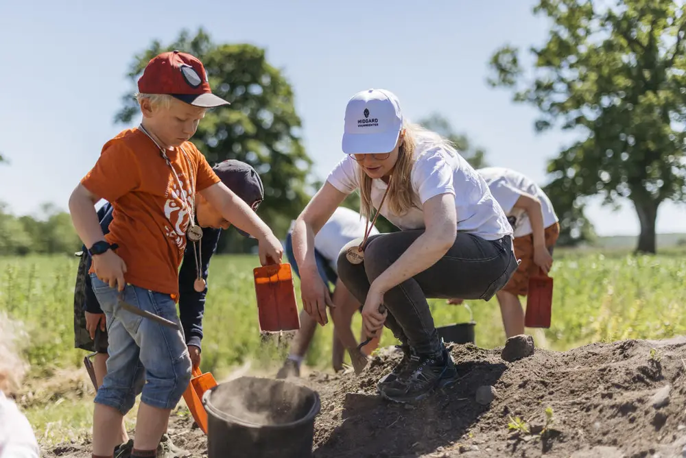 Et barn leker i sanda og en voksen sitter på huk og hjelper til. det er sol og sommer