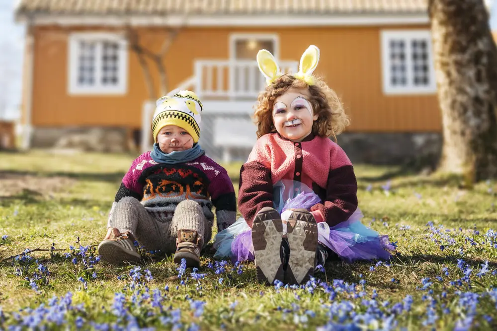 To små barn sitter foran et gult hus. De har påskehareører på seg. De sitter i gresset med blå blomster.