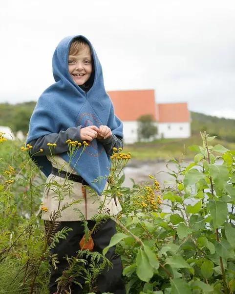 En gutt i vikingklær omgitt av blomster. En hvit kirkebygning med rødt tak i bakgrunnen.