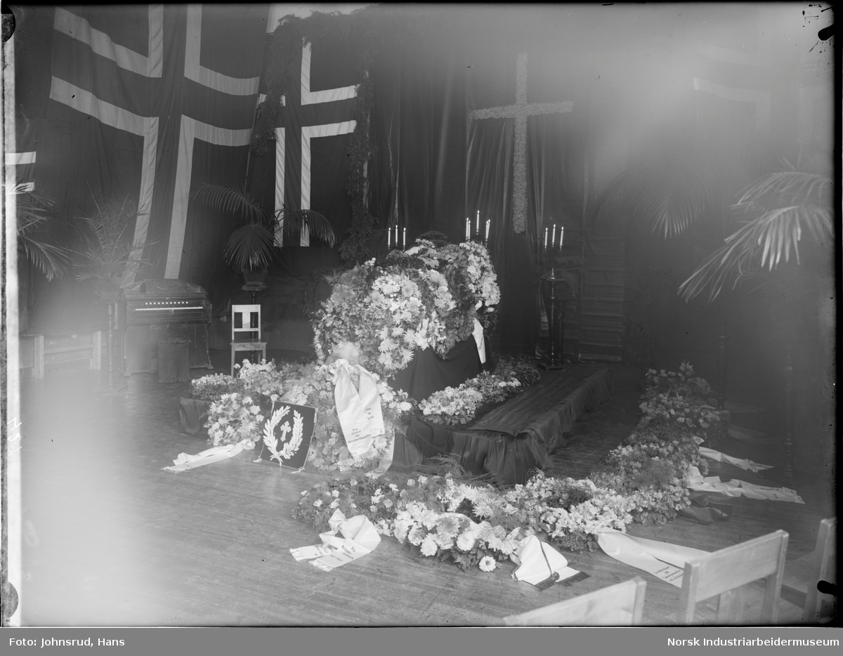 Begravelse til skolebestyrer Evje. Kiste omringet av blomster og blomsterkranser med tekstsløyfer. Norske flagg henger på veggene.