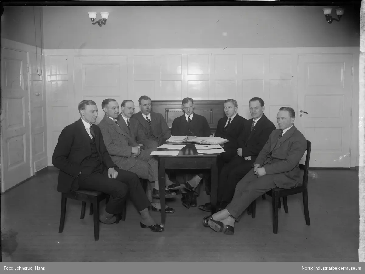Snøggs hovedstyre, åtte menn, sittende rundt et bord dekket med dokumenter. I bakgrunnen står ett piano. Alle mennene er kledd i dress og har gamasjer over skoene.