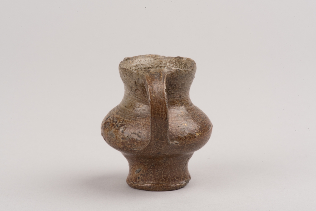 Ett så kallat salvekrus, ett miniatyrkrus av keramik, troligen stengods.
Runt, bukigt krus som smalnar av mot fot och mynning. Från mynning och ned till den bukigaste delen på kruset sitter ett öra.