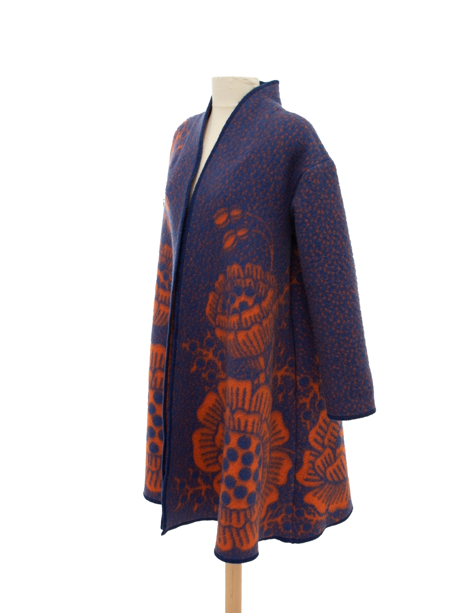 A-formet kåpe i blåfarget ull, dekorert med store orange blomster. Den har ståkrage, lav skuldersøm og sidelommer.