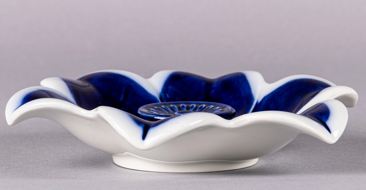 Askfat av flintgods, formgiven av Arthur Percy för Gefle Porslinsfabrik. Vitglaserad med flytande blå glasyr. Blomformad, sexflikig med upphöjd pistill i mitten. Mjukt uppsvängda blomblad och rund fotring. Tillverkningsår oklart men troligen 1930-tal.