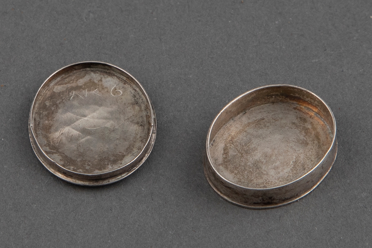 Liten oval eske i sølv med løst lokk. På lokket er det et speilmonogram.