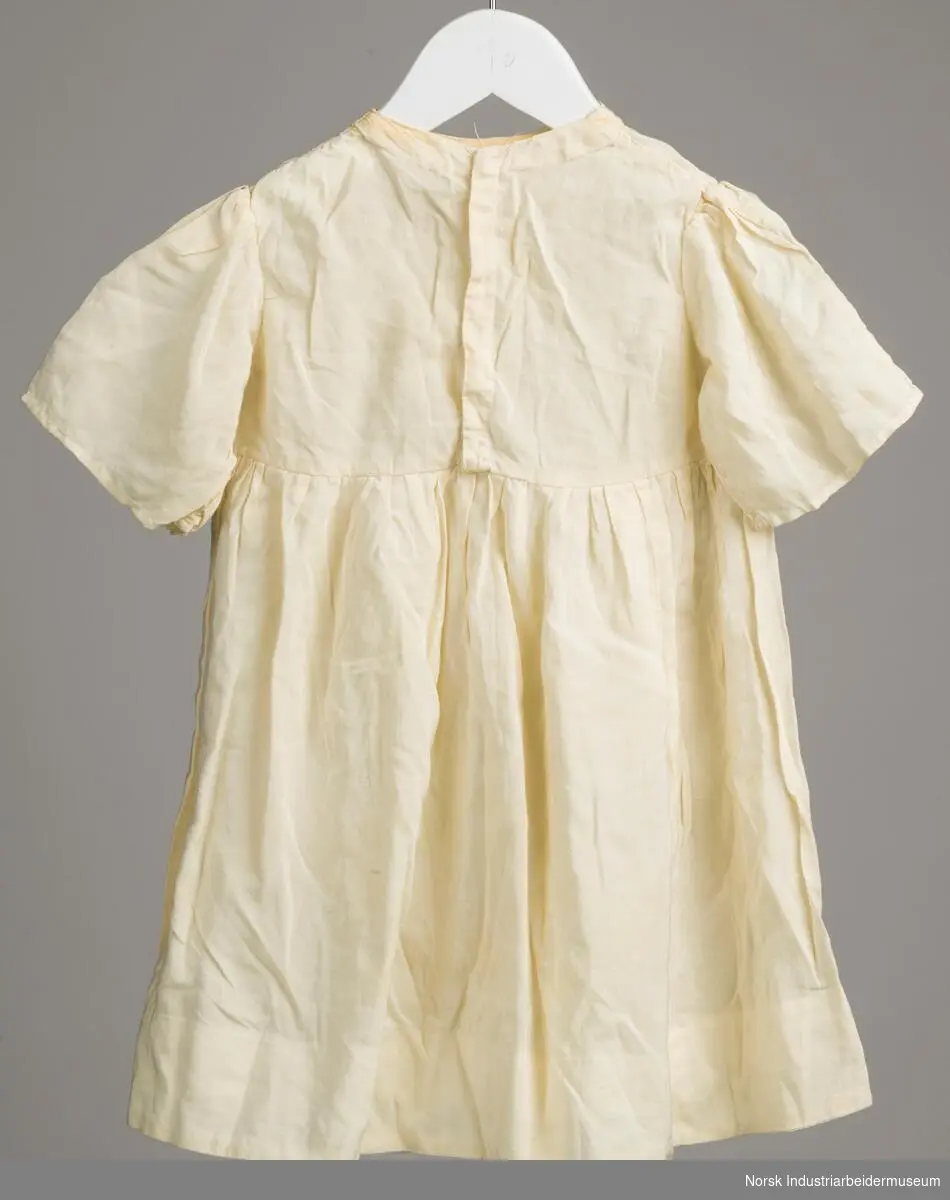 Hvit kjole i tynt stoff til barn. Åpning fra hals til liv lukkes med fire trykknapper.