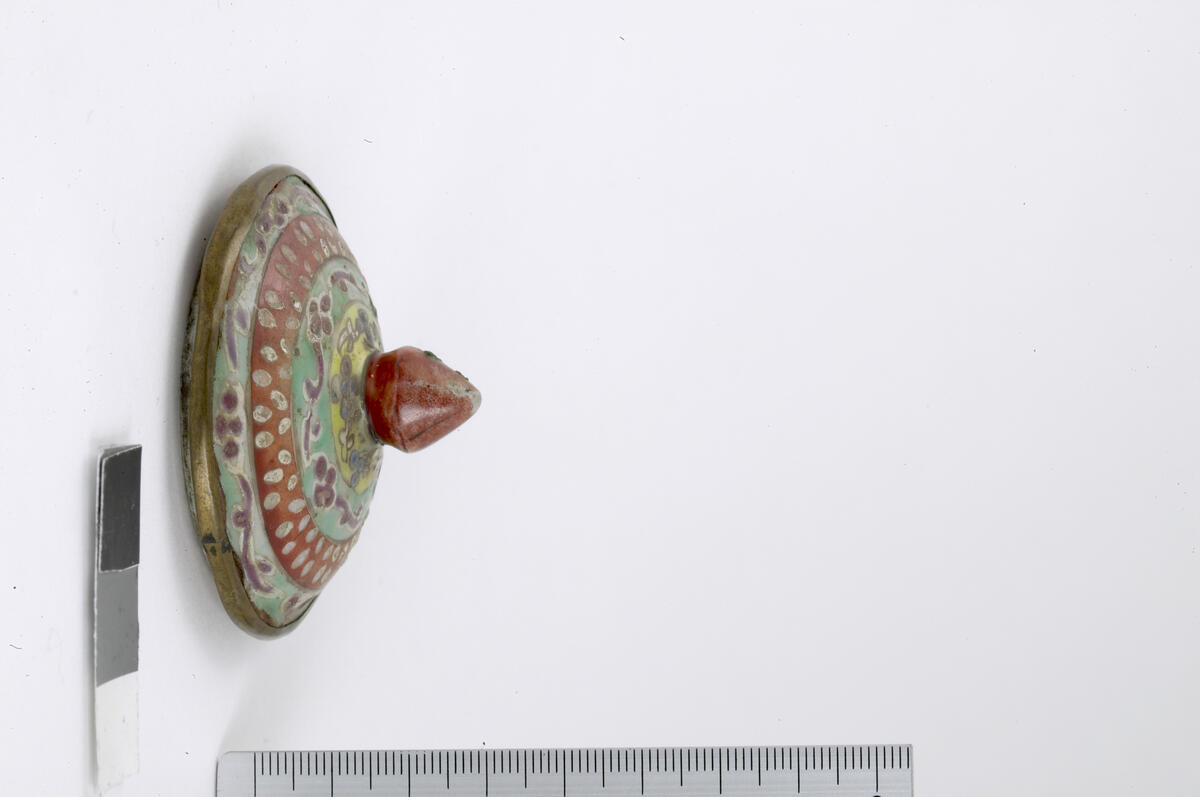 Bencharong. Small jar with cover. Bulbous, squat body. Two images of the deity Theppanom sitting in a triangular red medallion. Black background with the flame-pattern kranok in pink and white. Green band around the base, the enamel on the band around the shoulder is abraded off. The cover has four decorated bands circling the red lotus bud knob. Metal fitting on the footring, rim and cover rim. Cover height 3.6 cm, diameter 6.1 cm. Late 18th-19th century. 