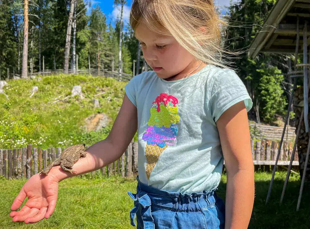 En jente med lyst hår og en blå T-skjorte ser ned på armen sin, på armen ligger en padde eller frosk, jenta står ved siden av en hytte