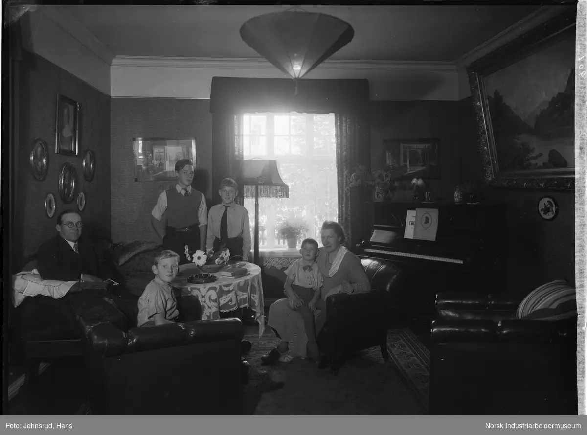Disponent Bøyesen med kone og fire sønner i stue. Familien sitter rundt stuebord, en av sønnene sitter på sin mors fang. Piano i hjørnet av stuen, bilder og maleri på veggene.