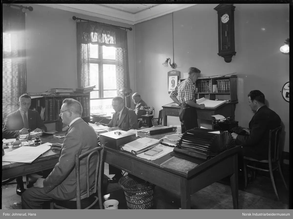 Kontorlokale til Kongsberg Sparebank. Seks menn i arbeid på kontoret. En arbeider på skrivemaskin, de resterende med protokoller eller papirer. Telefon stående på kontorpult. Spyttebakke under en av kontorpultene.