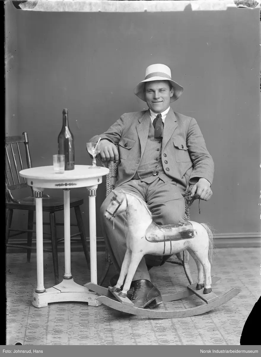 Portrett. Mann sittende i kurvstol i dress og hatt. Bord med flasker og glass ved siden av. Gyngehest foran.
