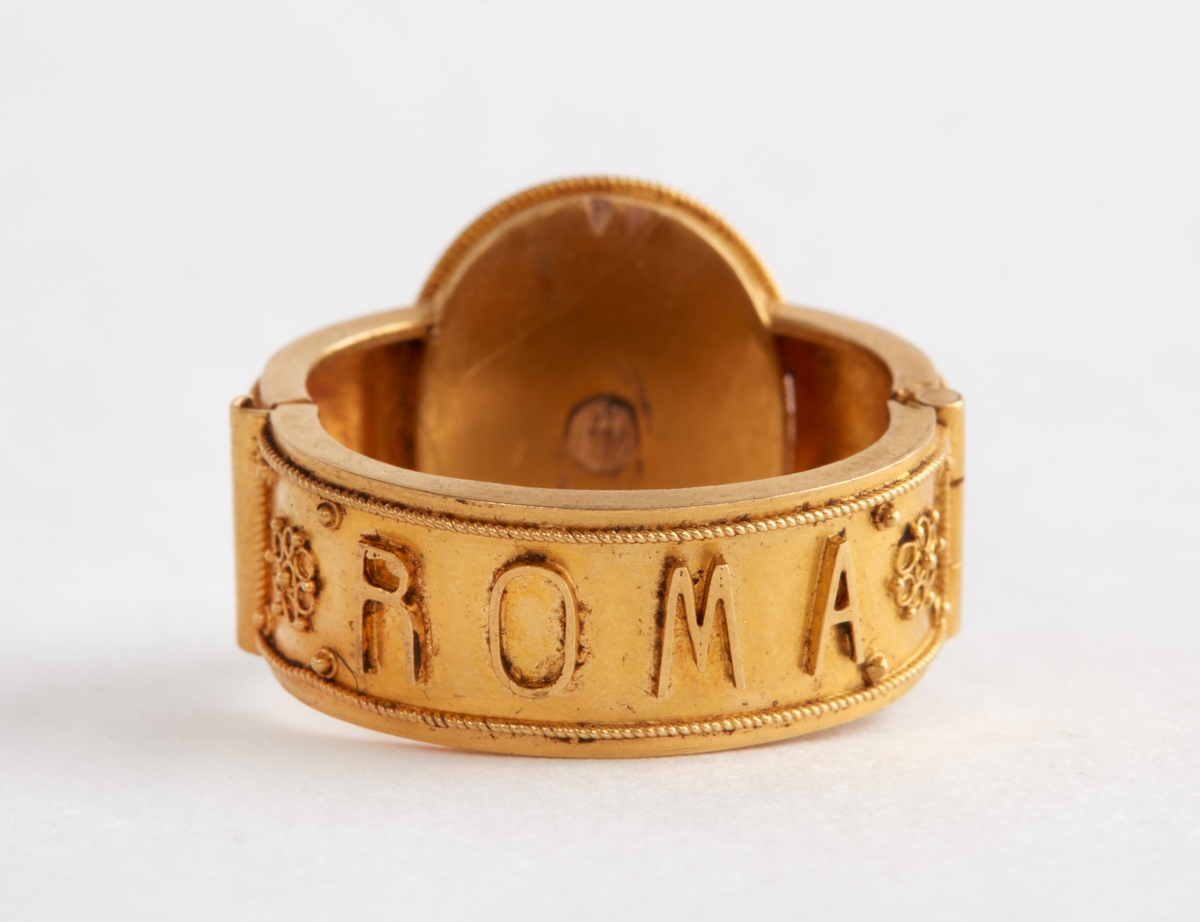 Ring av gul metall med mosaikinläggning. Med inskription: "ROMA".

Enligt uppgift från 1860-talet.