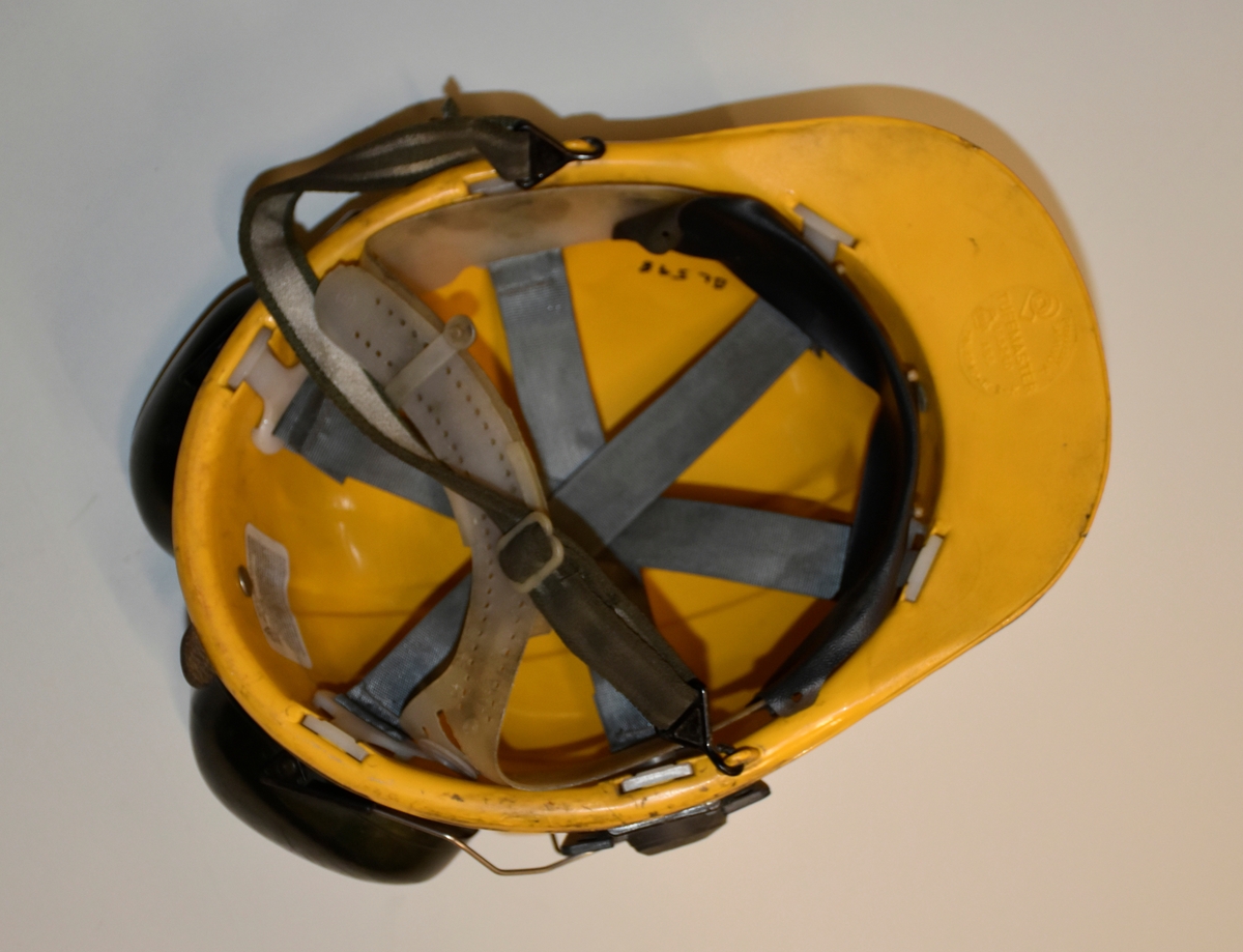 Hjelm i gul plast med fester i metall for lys foran og ledning til batteri bak. Påmontert hørselvern. Innmaten er laget av plast og nylonreimer. Hakerem.