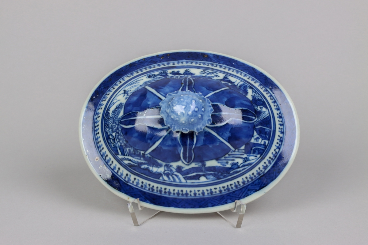 Oval shaped and slightly domed with knob in form of a flower head. On the top of lotus leaves and a list of pagoda lanscapes. The edge of the lid decorated with a dark blue  border in a criss cross pattern. All decor in blue underglaze. The inside of the lid without decor.