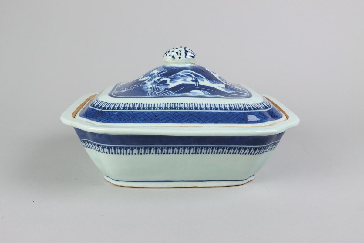 Square form with chamfered corners. The lid with a slightly domed form, on top a knob in form of a fruit. On the lid landscape scenes of pagodas, buildings,  bridges, figures with parasolls, gardens and waters. The edge is decorated with a wide dark blue border with a criss cross pattern. The sides of the dish are below the rim decorated with a criss cross patterned bord. All decor in blue underglaze. The base is not decorated.