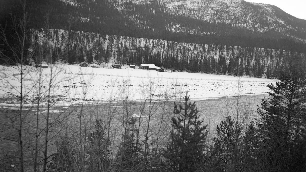Elvelandskap vinterstid, antakelig fra Glomma i Midt-Østerdalen en gang i 1930-åra.  Fotografiet er tatt fra en forholdsvis bratt elvebakke med furu- og bjørketrær tvers over elveløpet.  Elve går åpen, men har lav vannstand, og de steinete strendene på motsatt side av elva er fortsatt delvis snødekte.  Sletta ut mot elva er oppdyrket og inngjerdet.  Innerst på denne sletta, mot en bakenforliggende morenerygg med glissen furuskog, ligger et gardstun.  Bakenfor moreneryggen ses ei rand med tettere skog, og bakenfor denne igjen et fjellmassiv med mer glissen vegetasjon. 