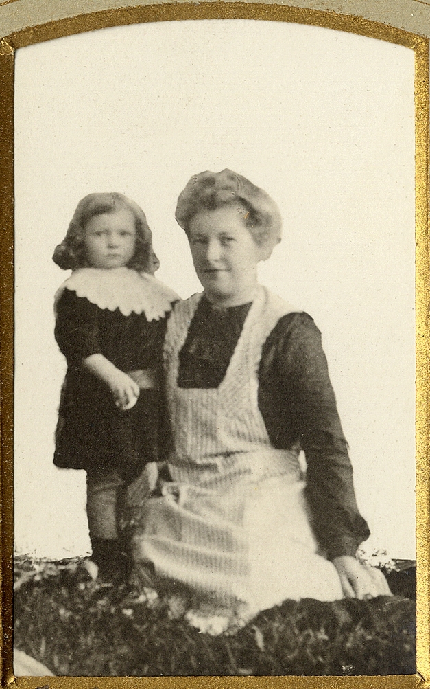 En kvinna i mörk klänning med förkläde poserar med ett litet barn hos fotografen.
Helfigur. Ateljéfoto.