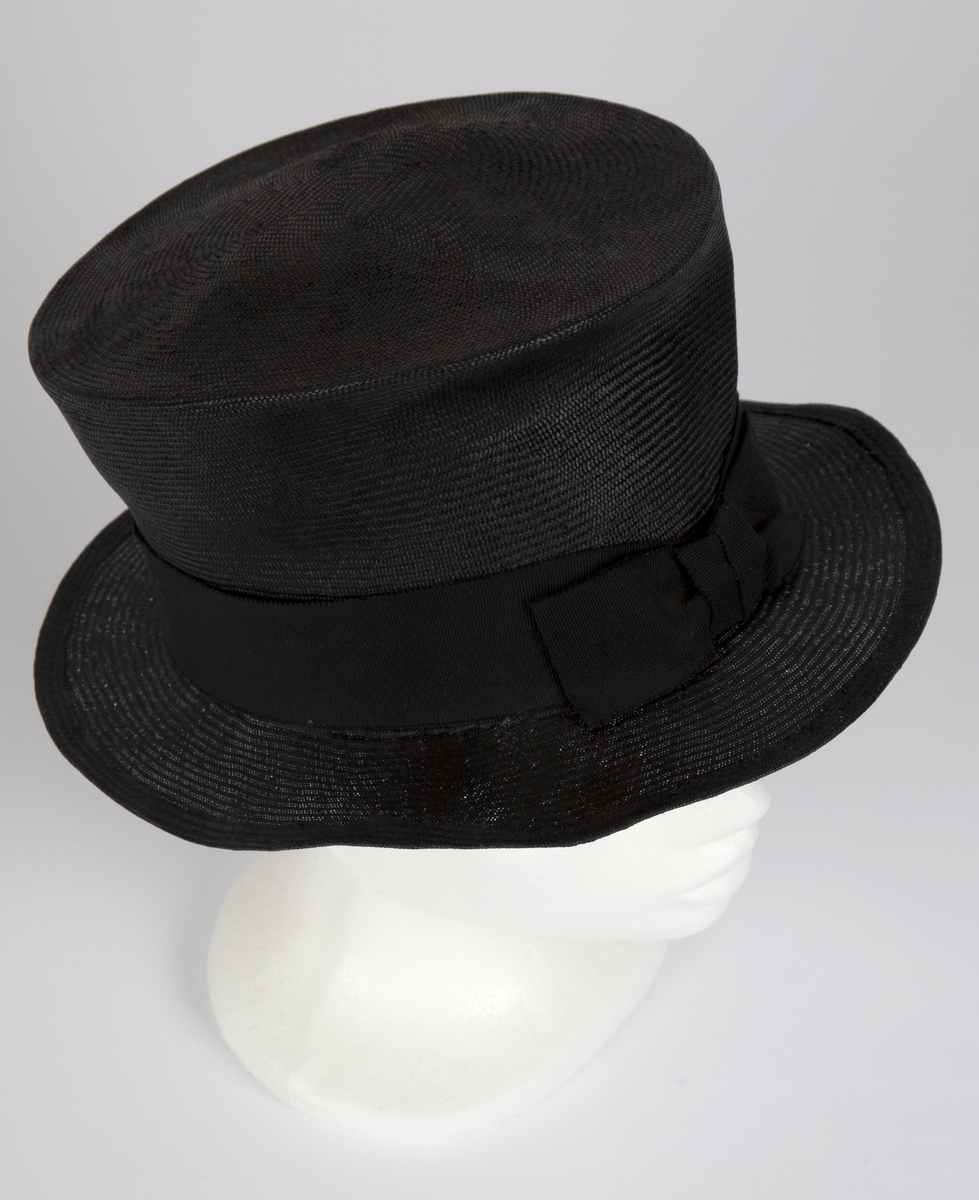 Svart, relativt høy "flosshatt", sydd av smale bånd av flettet strå. Sylindrisk, formpresset pull, som svinger ut i overkant, med påsydd, relativt smal brem, som vipper svakt oppover. Bremmens ytterkant er faldet, med smal oppbrett. Omkring pullens nederkant er hatten dekorert med et 4 cm bredt, ripsartet hattebånd prydet med en flat sløyfe av båndet midt foran. Pullens topp er svakt buet opp på midten, men likevel fremstår ytterkanten som en noe høyere rand rundt en liten høyde på midten. Hatten er ufôret, men har et 2,5 cm ripsbånd som svettebånd.