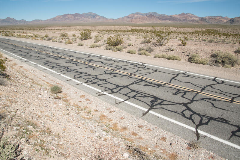 Motivet viser en vei i et ørkenlandskap med fjellkjeder i bakgrunnen. Veien er lappet på kryss og tvers med asfalt.