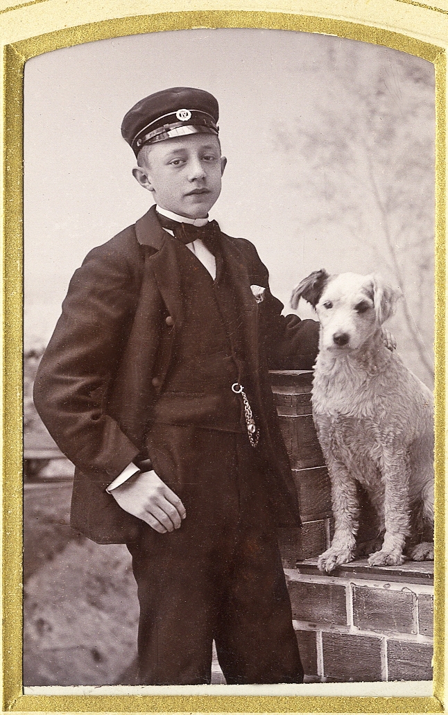 En pojke i kostym med stärkkrage, fluga och skolmössa. Han poserar tillsammans med en raggig hund hos
fotografen.
Knäbild. Ateljéfoto.