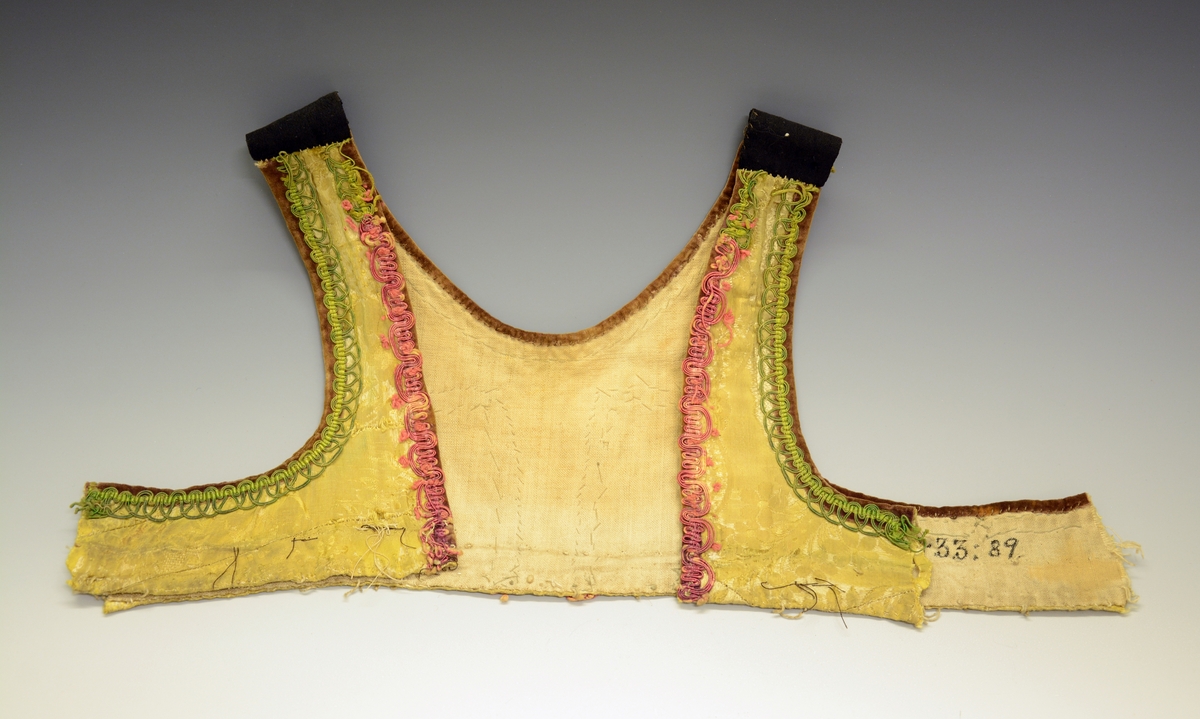 Ovaliv (upplut) av gul silkedamask med bordur av raude og grøne agramanur, Brun fløyelskant og lereftsfor. Ved ermstaupet er livet avklypt.  Fra protokoll.