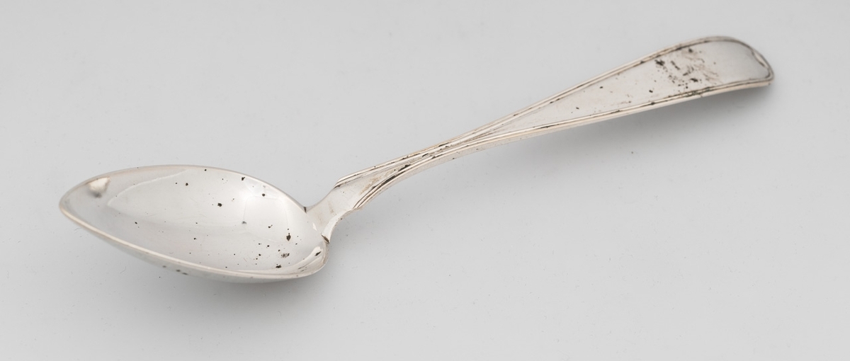 Skje i sølv med dråpeformet blad med spiss ende. Skaftet er buet med rille rundt kanten, samt avrundet avslutning.

