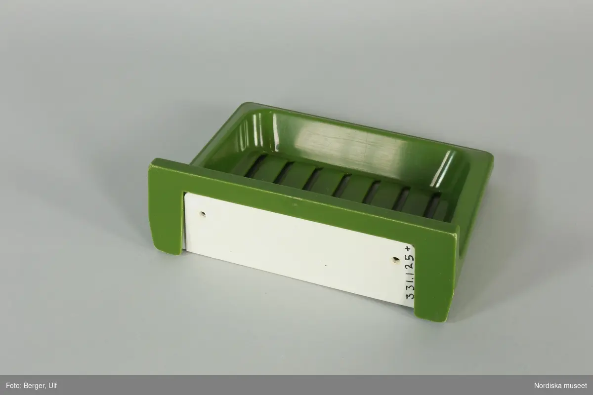 Tvålhållare, av grön (avocadogrön) formgjuten plast.  Löst vitt bakstycke/monteringsplatta med text "OBEN" och en pil. 
/Johan Åkerlund 2010
