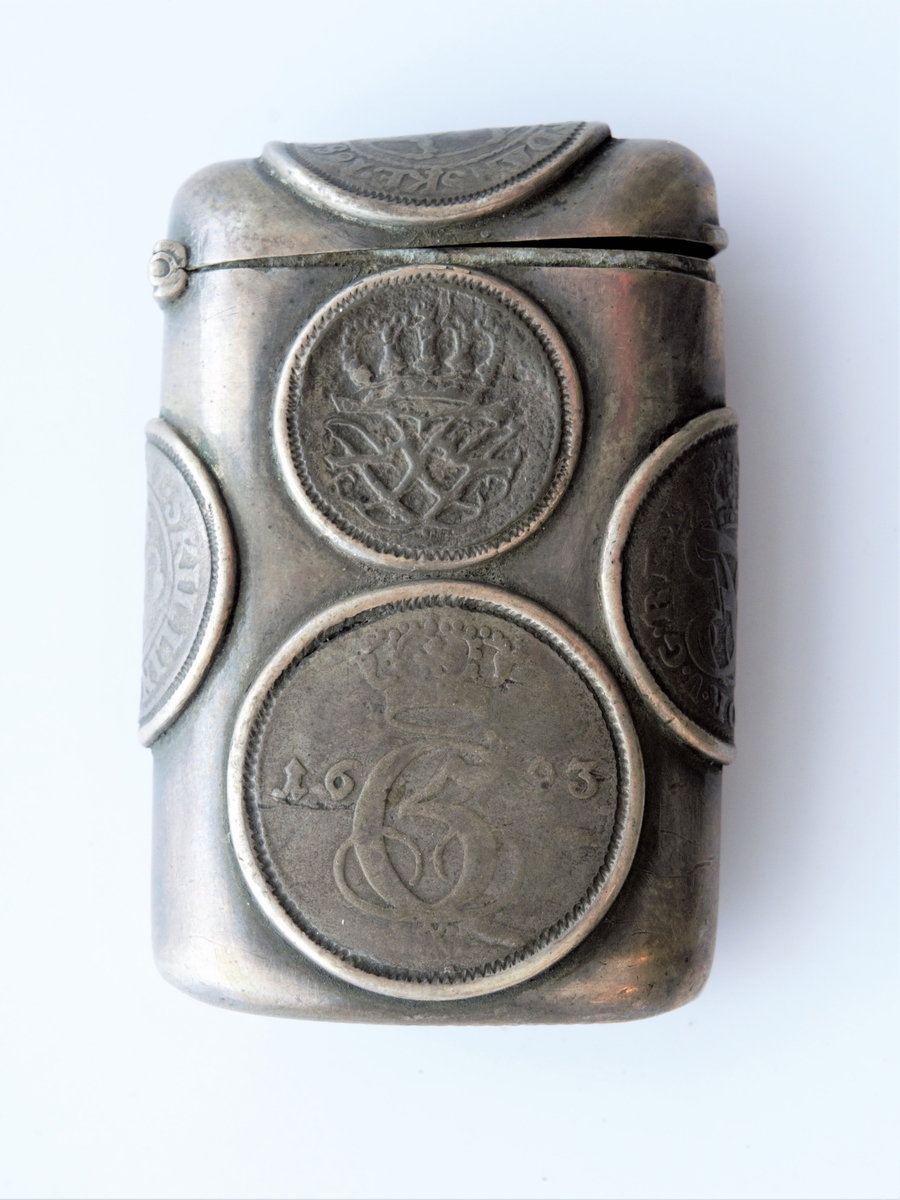 Beholder med ripeflate; brukt til svovelstikker. Innfeldt 6 stk dansk-noske mynter utvendig. Disse er datert fra 1689, 1693, 1709 m.v.