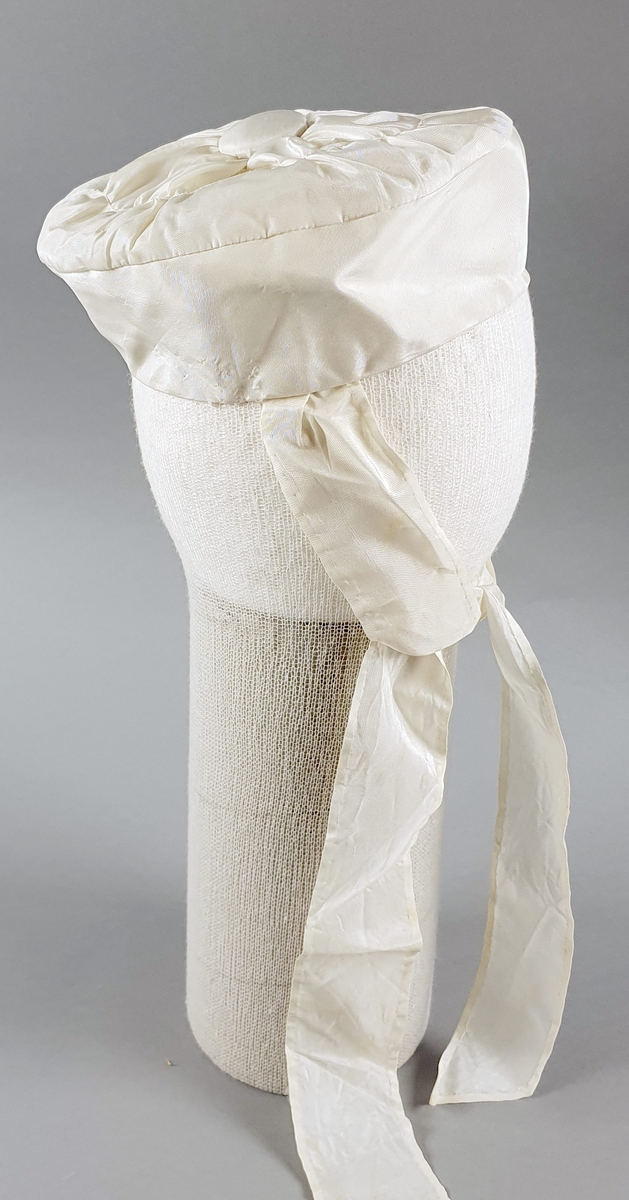 Hvit babylue av silke med lange bånd til å knyte under haken. Stor knapp på toppen og bånd rundt kanten.