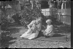 Kvinne og barn sittende på teppe i hage. Kvinnen har en hund
