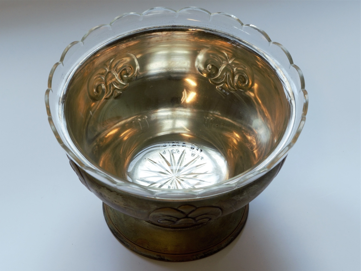 Skålformet potteskjuler i to deler. Den ytre delen er av sølvplett og den indre er av glass. Amerikansk design. Presset blomstermønster langs kanten av sølvskålen. Glasskålen har slipt stjernemønster i bunnen og dekorativ bord langs kanten.