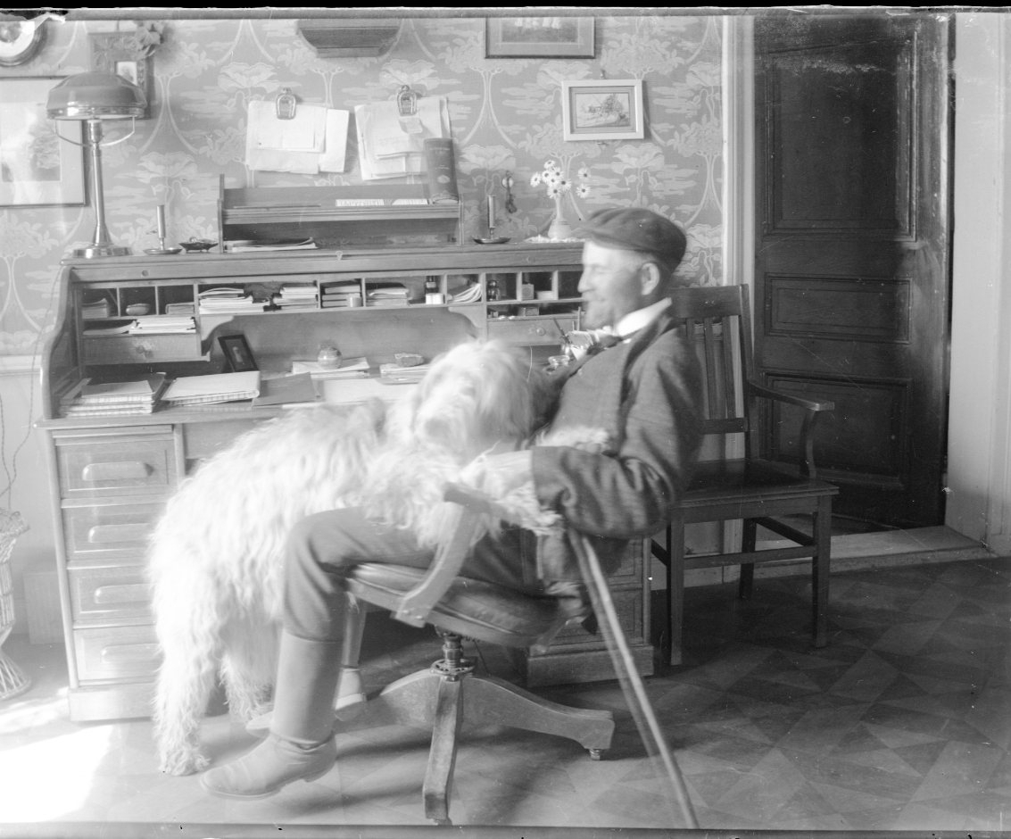 En man med keps sitter tillbakalutad i en kontorsstol vid ett skrivbord med en stor lurvig hund i knät.