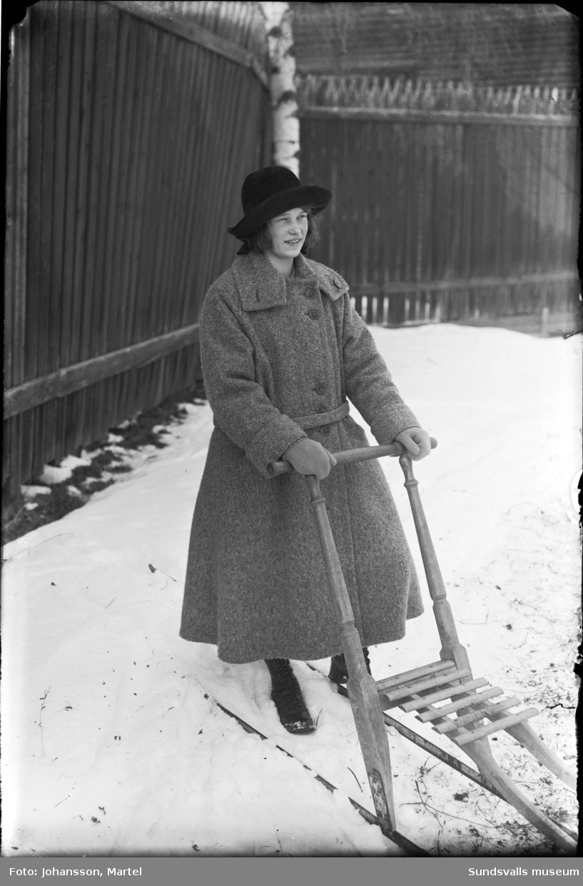 Anna Viklund, Sörfors, med vinterkappa, hatt och spark. Senare gifte hon sig Lockner och var bosatt på Alnö. I bakgrunden ett högt staket.