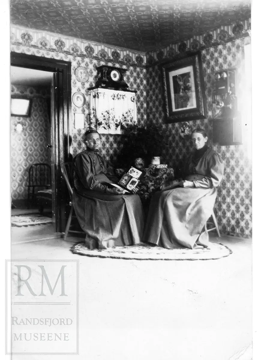 To eldre damer sitter ved et lite hjørnebord og blar i album. Telefon på veggen til høyre.