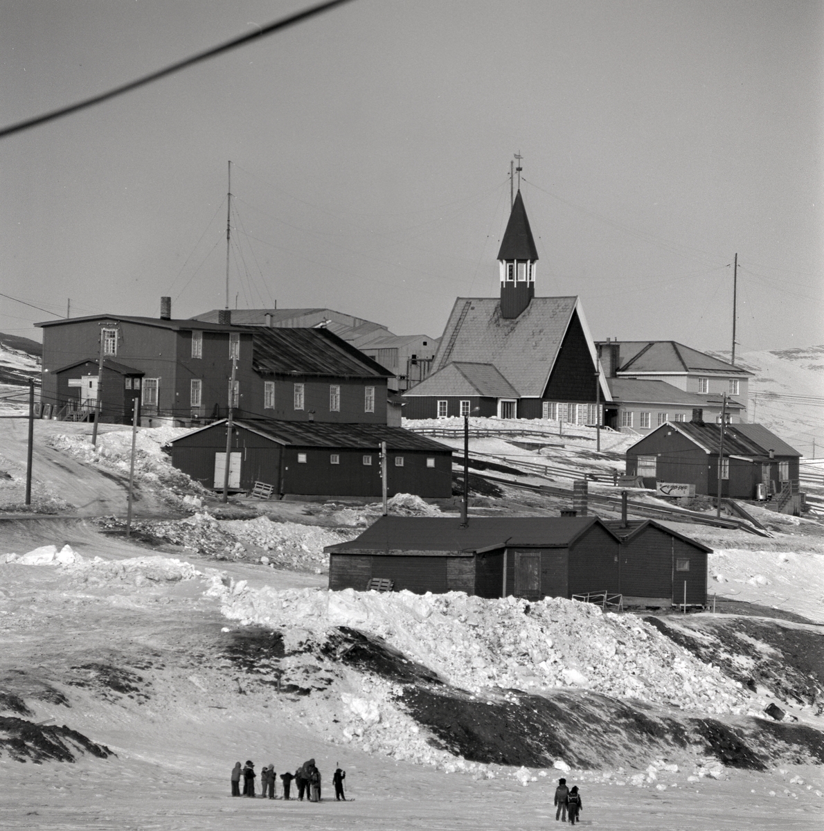 Barn som lekerog  går på ski nedenfor fjøset og bakeriet i Gamle Longyearbyen. Gamle sykehus midt i bildet, Svalbard kirke, mellageret, taubanesentralen.