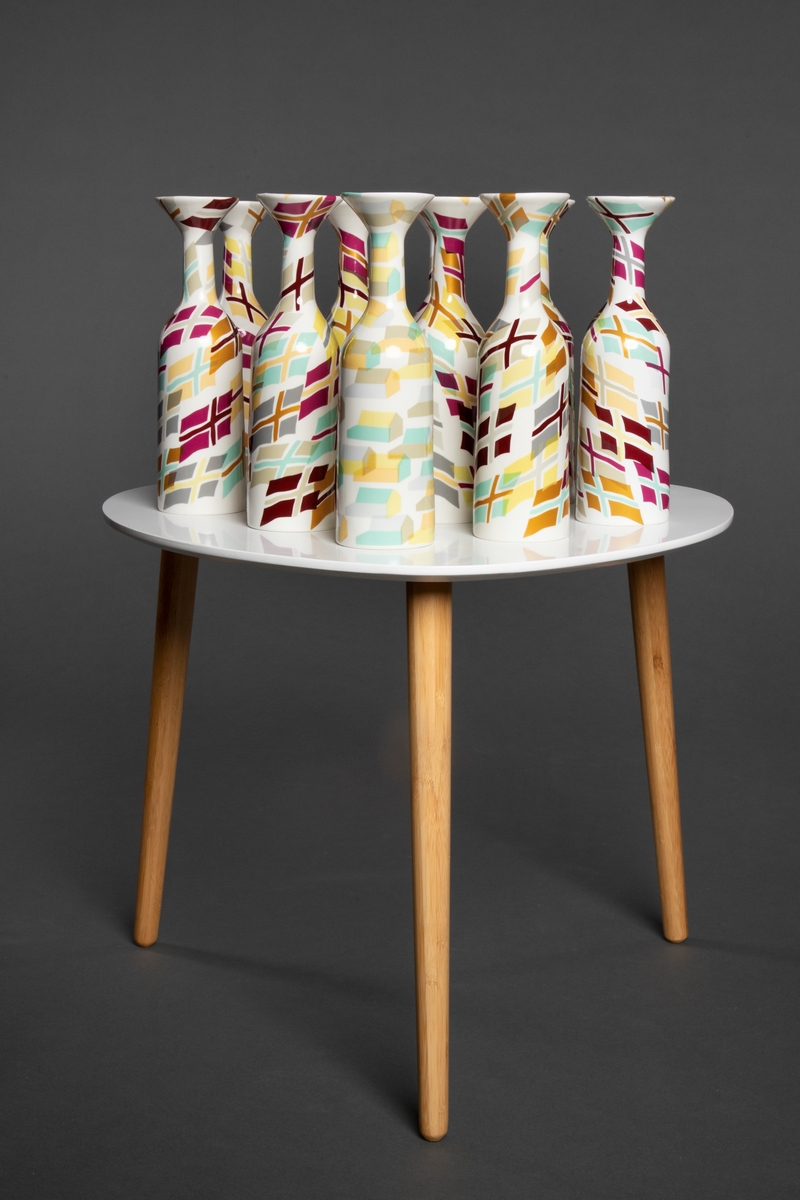 11 porselensvaser på et hvitt bord med tre trebein. Vasene er dekorkert med dekaler som viser variasjoner over det samme flagget. En av vasene har hus som motiv.