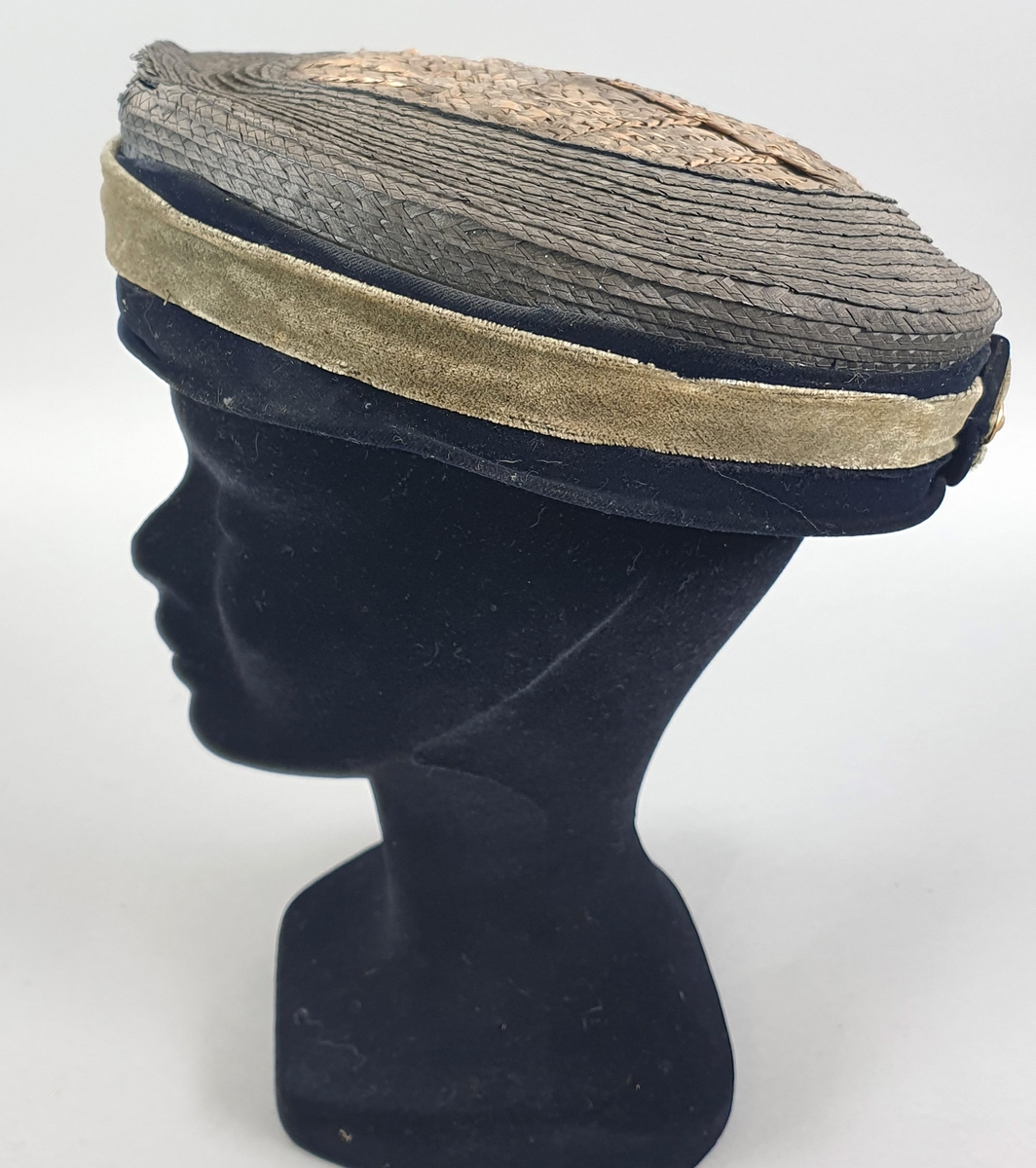 Rund og flat stråhatt med kant av grønn og blå fløyel og gul fløyelssløyfe midt bak. Kant på innsiden av skinn, som er løsnet fra hatten.