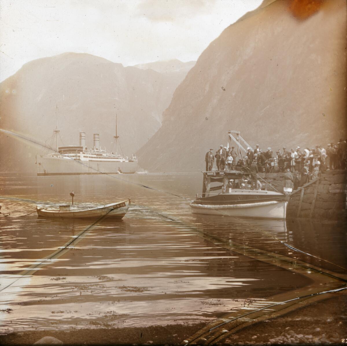 Dias. Passasjerskipet "Stavangerfjord" ligger på fjorden ved Hellesylt. En stor gruppe kvinner og menn står på en brygge. I forgrunnen ligger to mindre båter på vannet. 