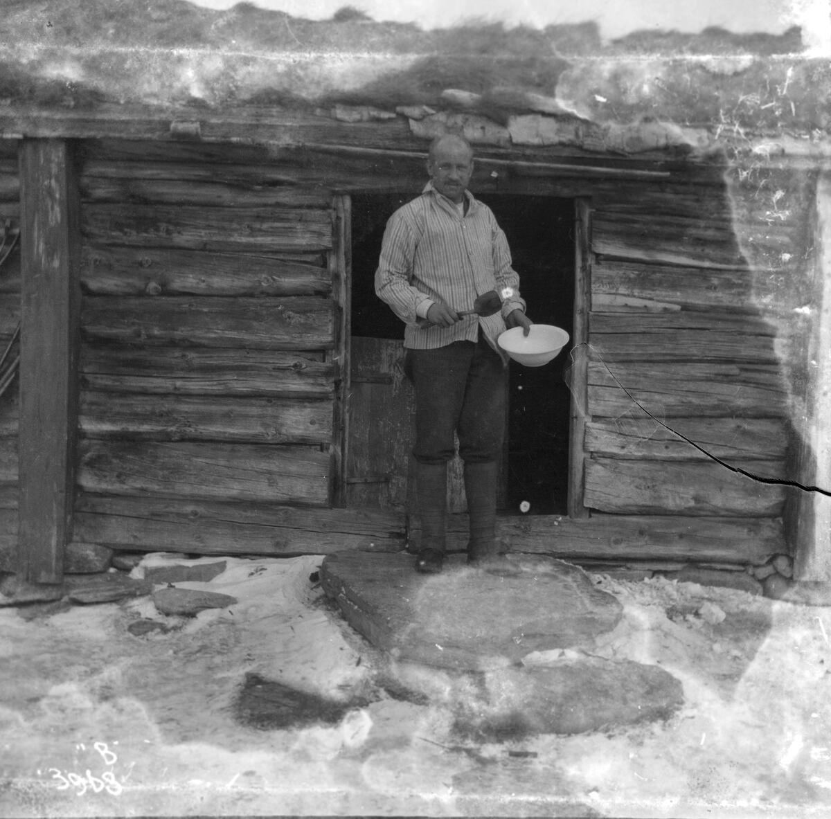 En mann står utenfor en laftet tømmerhytte eller et seterhus med en bolle i hånden.