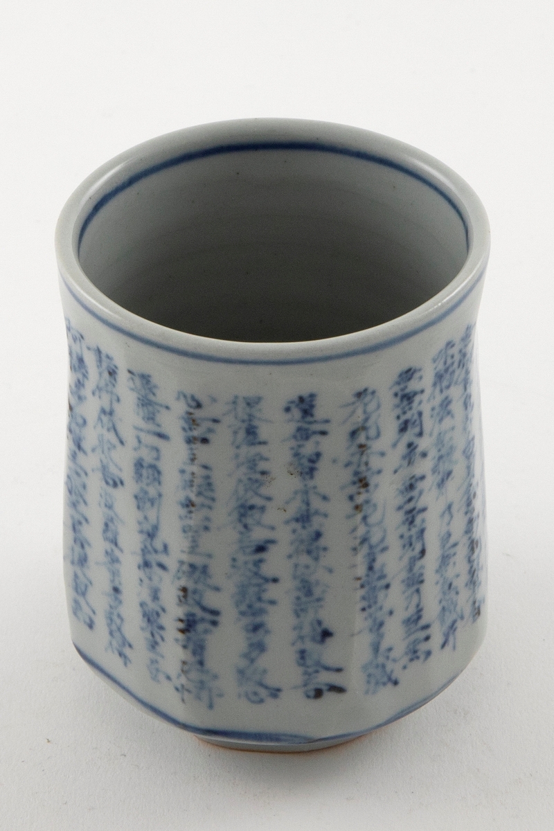 Tekopp i keramikk. Sylindrisk form med liten fot. Glasert med blå dekor som viser skrifttegn samt en blå sirkel rundt koppen ved øvre kant, nedre kant og rundt foten.