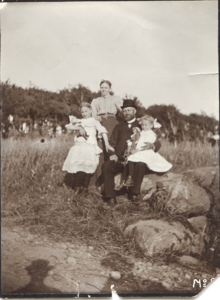 Familjen Gustafsson sitter på några stenar framför ängarna vid Kållered Stom 1:3 "Nygård" cirka 1920. Carolina Gustafsson (1873 - 1949, född Alm) från Ekan, Johan Gustafsson (1867 - 1956) från Högen samt systrarna Ingeborg (1901 - 1987, gift Johansson) och Astrid (1907 - 1994, gift Jägerström, Råda).