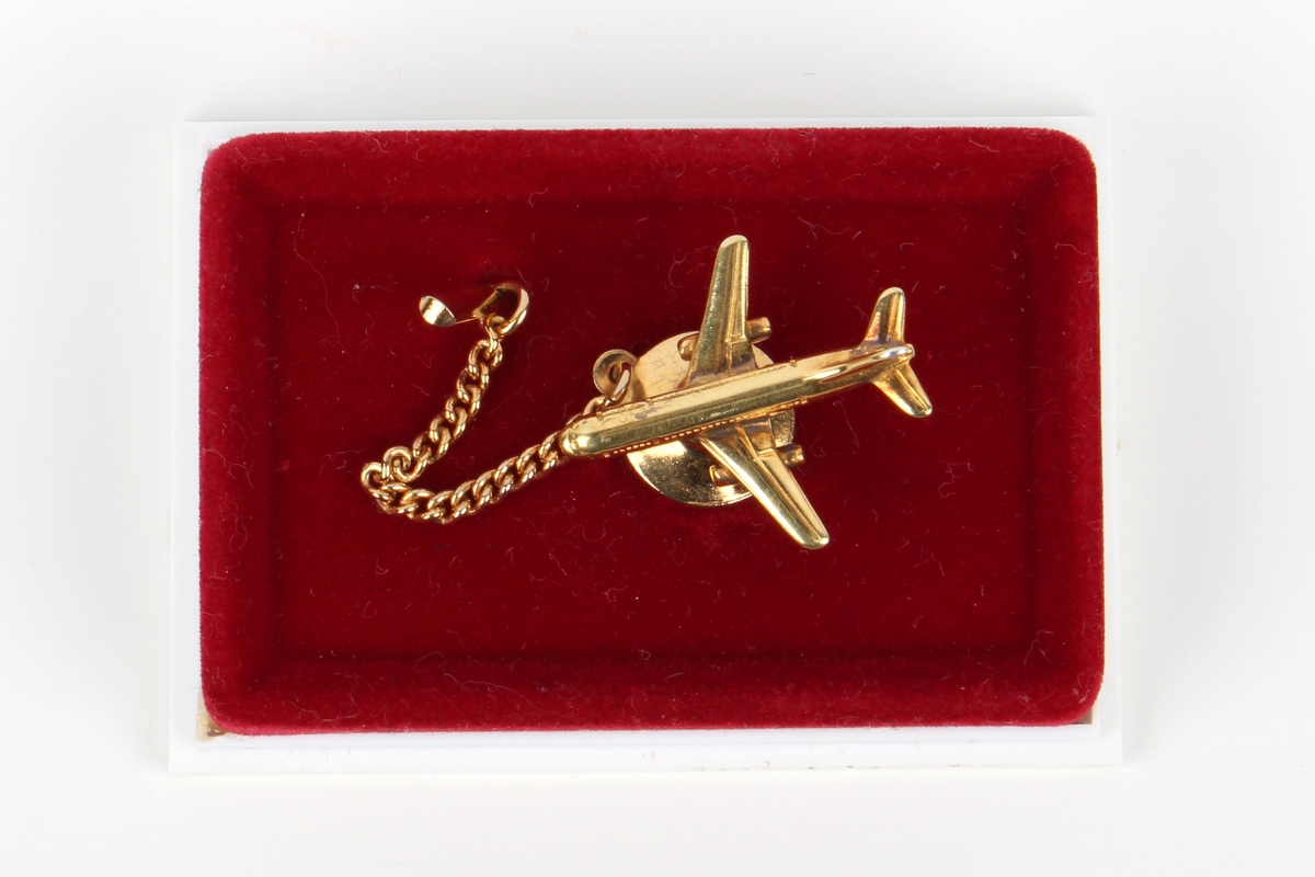 Gullfarget slipsnål med formet som et fly av modellen Boeing 737. Festet på et stykke rød kartong, ligger i lite etui av plast med hvit bunn og gjennomsiktig lokk. Lokket med gullfarget Braathens SAFE-logo.